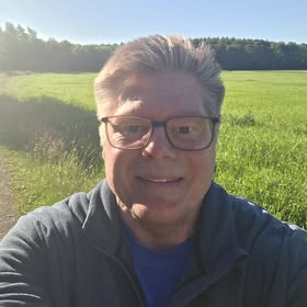 Leende äldre kvinna med glasögon tar selfie utomhus på grön äng i kvällsljus.